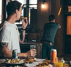 Man eating pizza watching someone throw an axe at a target