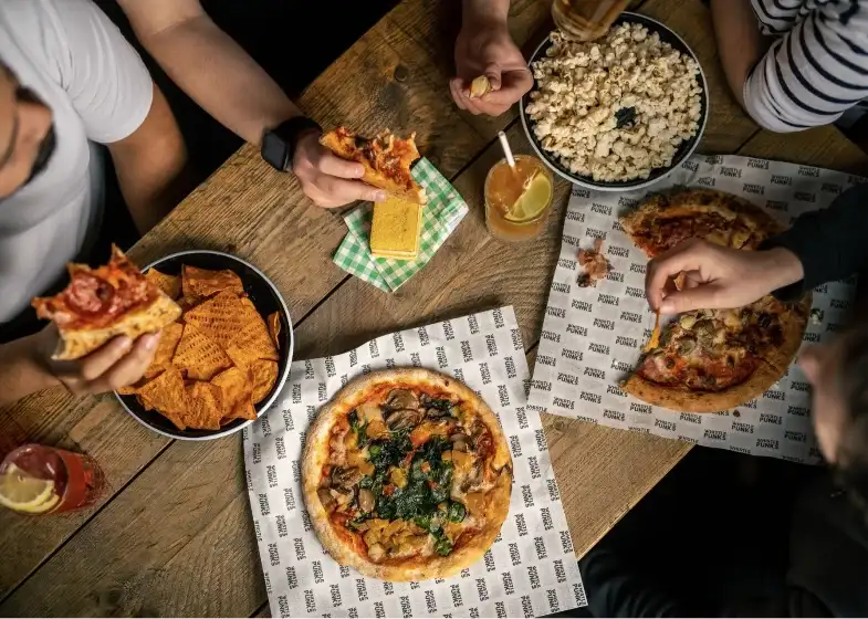 a birds eye view of a table with people sat around it eating pizza, popcorn and nachos.